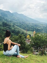 Side view of young woman sitting on mountain against cloudy sky