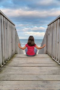 Rear view of girl sitting on pier against sea and sky