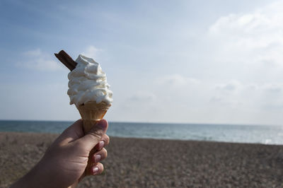Cropped hand holding ice cream at beach against sky