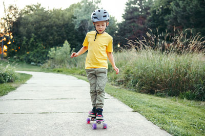 Happy caucasian boy in grey helmet riding skateboard on road in park on summer day