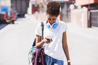 Smiling young woman wearing headphones looking at cell phone outdoors