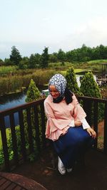 Woman in hijab sitting by railing against trees and lake