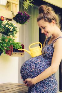 Pregnant woman standing with watering can on belly at balcony