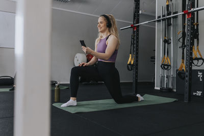 Young woman using phone while stretching on mat in gym