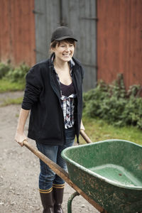 Happy female farmer with wheelbarrow against barn
