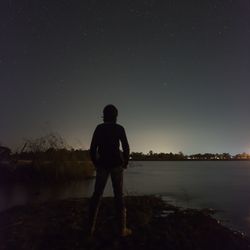Rear view of silhouette man standing by lake against sky at night
