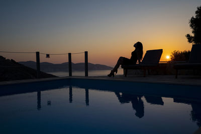 Silhouette of woman sitting on lounge chair by swimming pool against sky during sunset