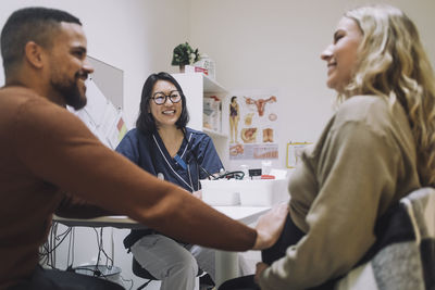 Happy female gynecologist looking at smiling man touching stomach of pregnant woman in clinic
