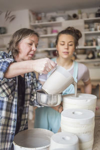 Young female employee looking at mature potter pouring clay in vase at workshop