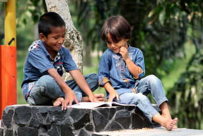Boys reading book while sitting on seat against tree