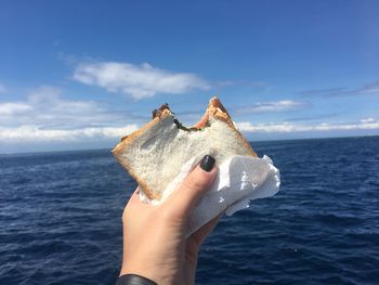 Close-up of hand holding sandwich by sea against blue sky