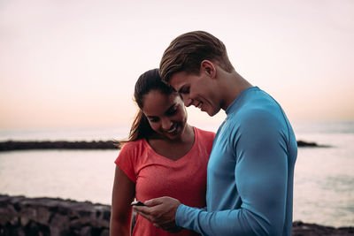 Smiling friends using mobile phone against sea during sunset