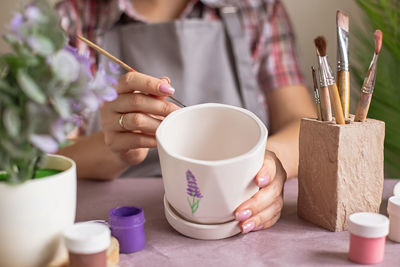 Women in gray apron paint a white flower ceramic pot on the table