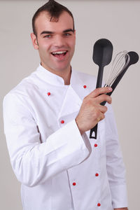 Portrait of happy chef holding kitchen utensils while standing against gray background