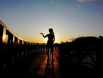 Silhouette girl gesturing while standing on bridge against sky during sunset