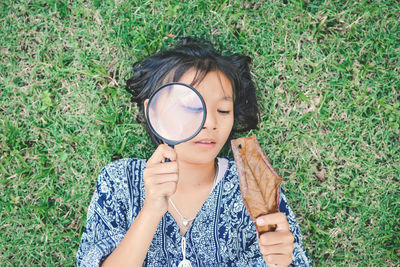 High angle view of girl holding magnifying glass on face while lying on grassy field