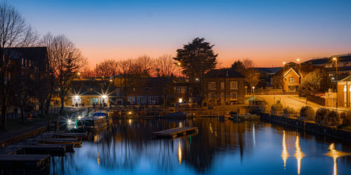 Illuminated buildings by river against sky at sunset