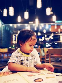 Cute baby girl looking at menu while sitting in illuminated restaurant