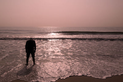 Rear view of silhouette standing on beach against sky during sunset