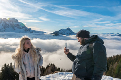 Husband using mobile phone, taking photos of wife in amazing mountains