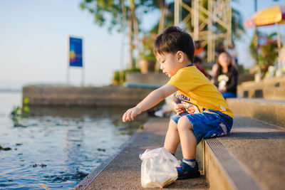 Side view of cute baby boy throwing food in lake while sitting on steps during sunset