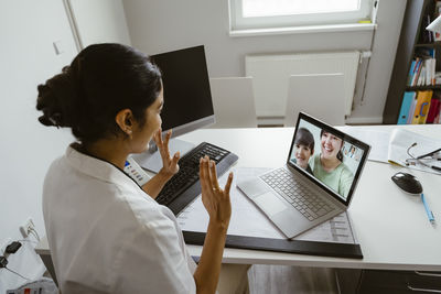 High angle view of female pediatrician waving patients over video call through laptop at desk in clinic