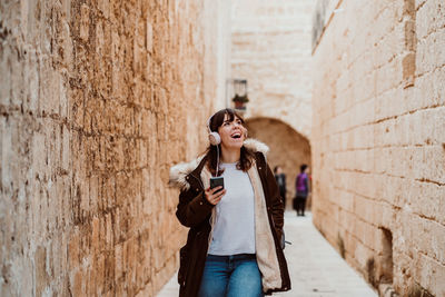 Young woman using phone while standing against wall