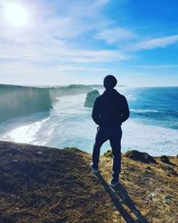 Rear view of man standing on cliff by sea against blue sky