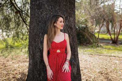 Smiling woman looking away while leaning on tree trunk in park