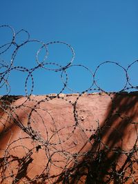 Low angle view of razor wires on surrounding wall against clear blue sky