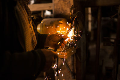 Unrecognizable elderly craftsman using bench grinder to shape metal part while working in dark workshop