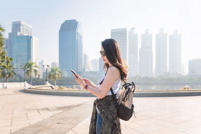 Side view of young woman using mobile phone against buildings in city