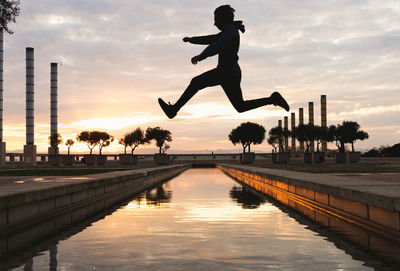 Full body of unrecognizable male silhouette jumping above lake while doing parkour trick on sundown