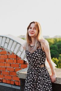 Portrait of beautiful young woman wearing sundress while standing on terrace against clear sky