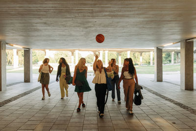 Teenage girl throwing basketball while walking with female friends under bridge
