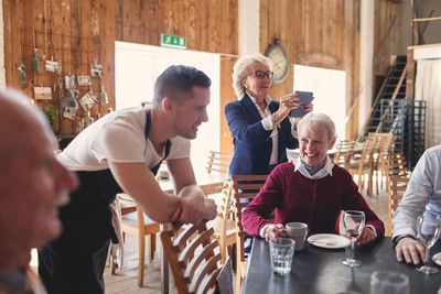 Young waiter leaning on chair while senior friends socializing in restaurant