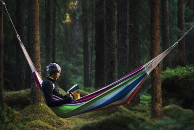 Man reading book while sitting in hammock at forest