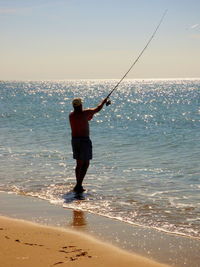 Full length rear view of shirtless man fishing while standing on shore