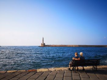 People sitting on bench looking at sea against clear sky