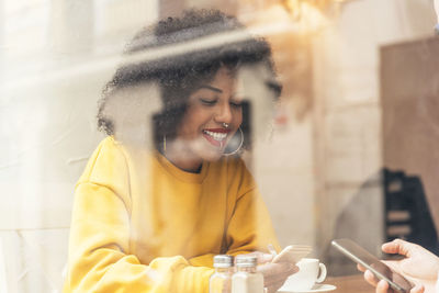 Smiling young woman using phone in cafe seen through window