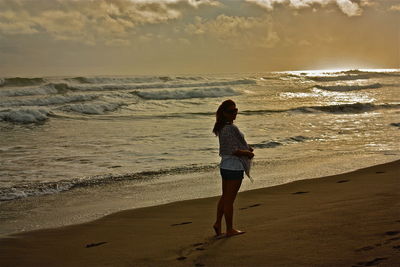 Full length of woman standing on shore at beach against sky during sunset