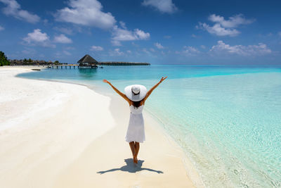 Rear view of mid adult woman with arms raised standing at beach against blue sky during sunny day