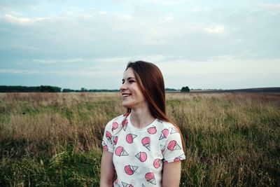 Happy beautiful woman looking away while standing on grassy field against cloudy sky