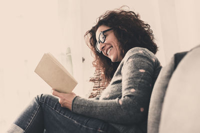 Smiling woman reading book while sitting on sofa at home