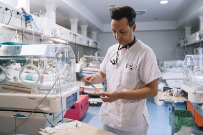 Pediatrician in white uniform filling syringe with vaccine for injection in room with incubators for newborns in hospital
