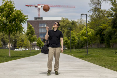 Full length of man playing with basketball while standing on footpath against sky