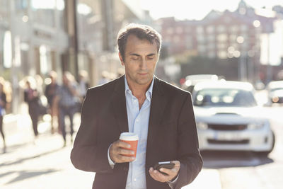 Businessman using mobile phone and holding disposable coffee cup while walking on city street