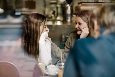 Smiling teenage female friends talking while sitting at cafe seen through glass window