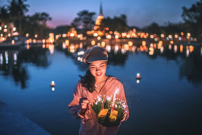 Young woman burning sparklers while standing by lake against sky at night