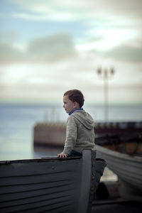 Cute boy in a gray knitted sweater sits on an old wooden boat in cloudy weather and looks away
